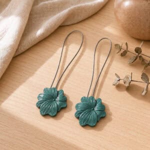 02 (pair, cozy styled): Two dark green flower tiebacks with looped silver cables resting on a warm wood surface beside soft linen and dried eucalyptus, styled to show the pair in a cozy neutral setting.