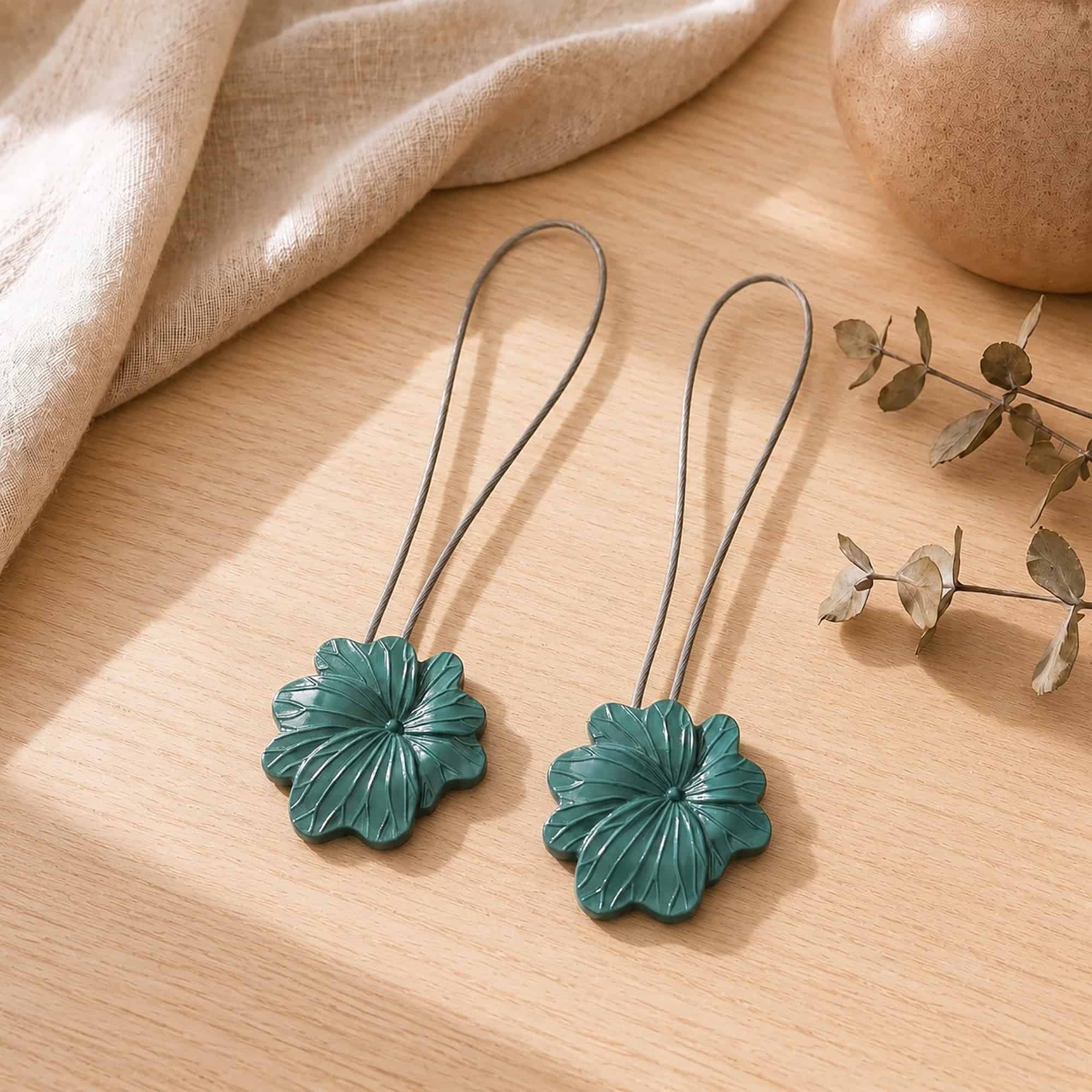 02 (pair, cozy styled): Two dark green flower tiebacks with looped silver cables resting on a warm wood surface beside soft linen and dried eucalyptus, styled to show the pair in a cozy neutral setting.