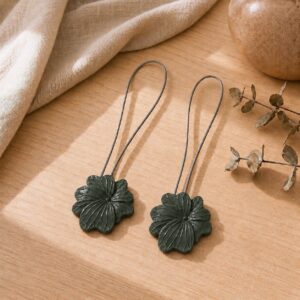 02 (pair, cozy styled): Two khaki flower tiebacks with looped silver cables resting on a warm wood surface beside soft linen and dried eucalyptus, styled to show the pair in a cozy neutral setting.