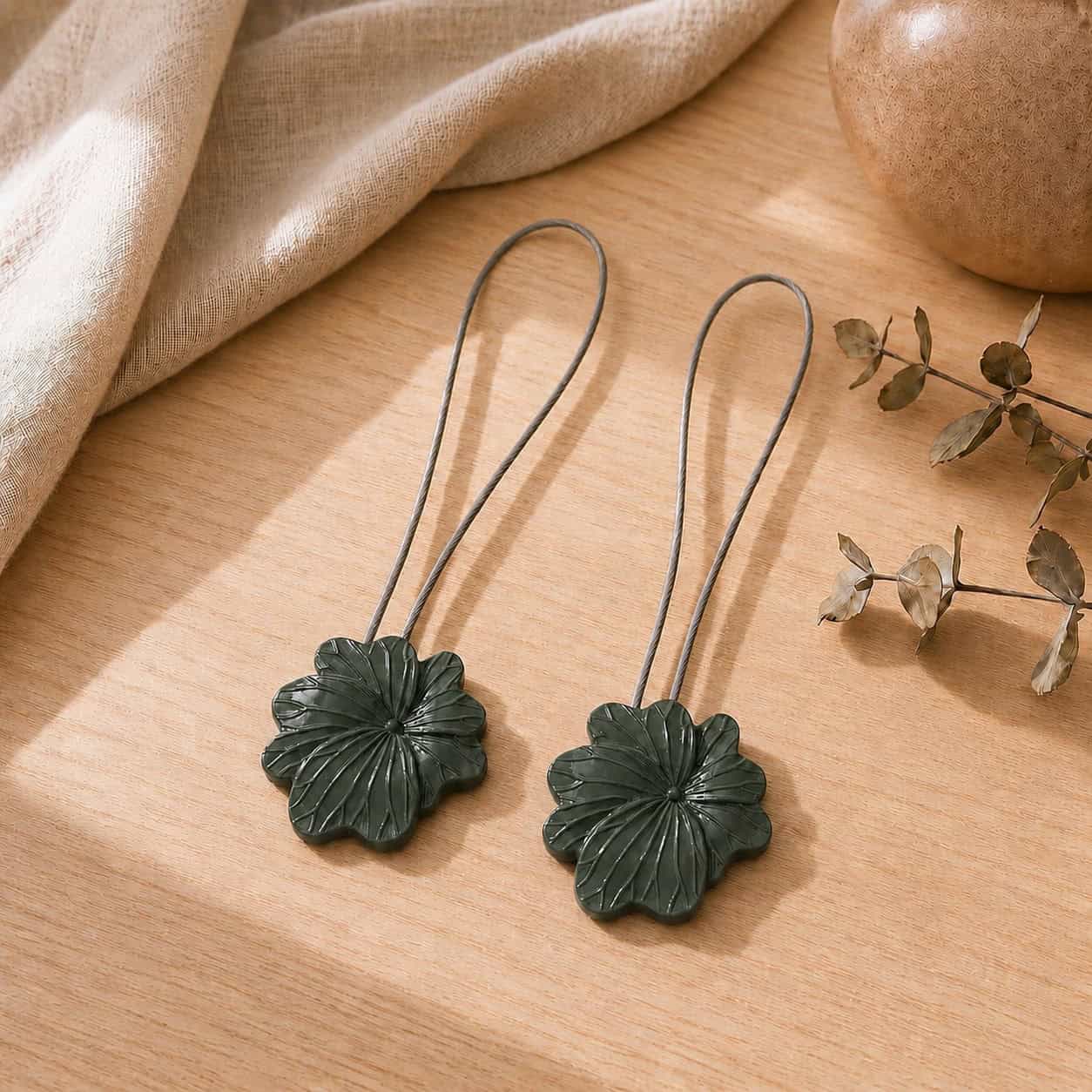 02 (pair, cozy styled): Two khaki flower tiebacks with looped silver cables resting on a warm wood surface beside soft linen and dried eucalyptus, styled to show the pair in a cozy neutral setting.