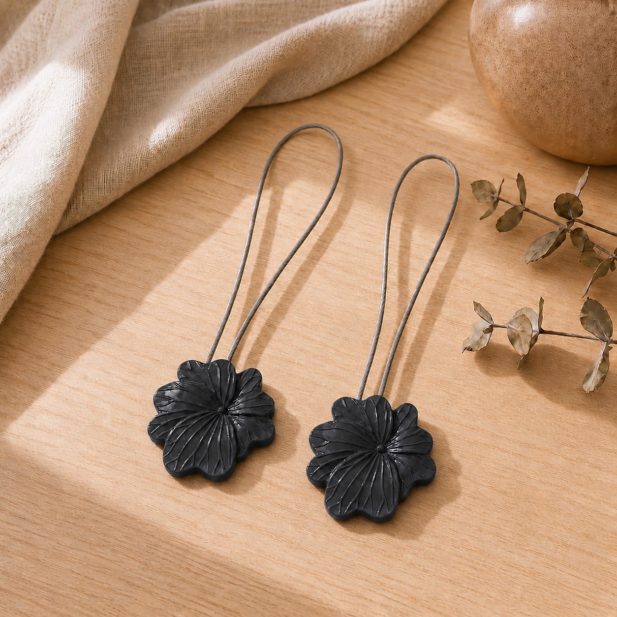 02 (pair, cozy styled): Two matte black flower tiebacks with looped silver cables resting on a warm wood surface beside soft linen and dried eucalyptus, styled to show the pair in a cozy neutral setting.