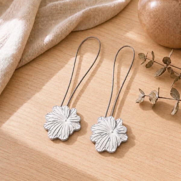 02 (pair, cozy styled): Two white flower tiebacks with looped silver cables resting on a warm wood surface beside soft linen and dried eucalyptus, styled to show the pair in a cozy neutral setting.