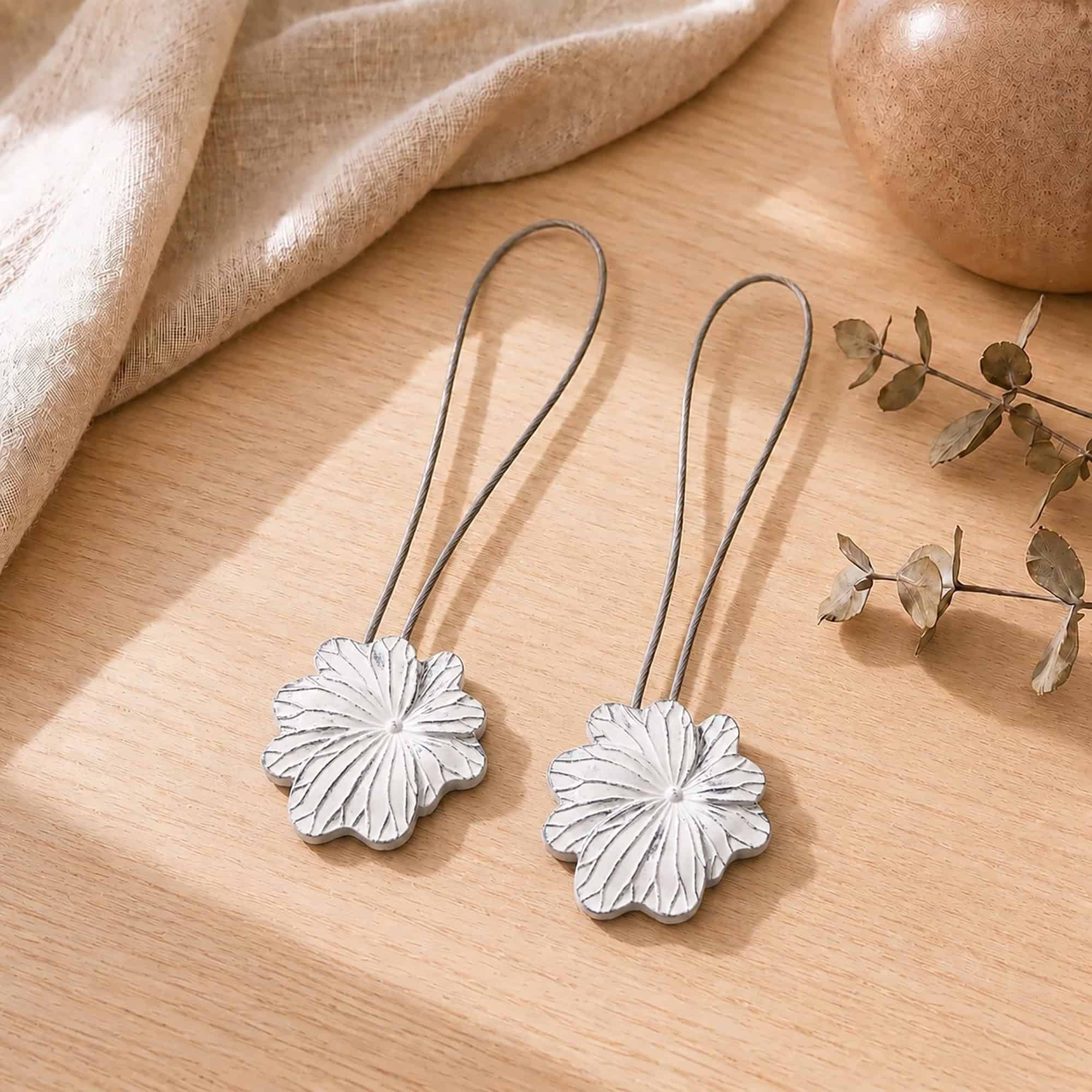 02 (pair, cozy styled): Two white flower tiebacks with looped silver cables resting on a warm wood surface beside soft linen and dried eucalyptus, styled to show the pair in a cozy neutral setting.