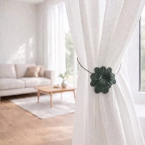 08 (living room lifestyle): Sheer white curtain held back by the khaki flower tieback in a bright living room with a neutral sofa and wood coffee table softly blurred behind.