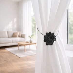08 (living room lifestyle): Sheer white curtain held back by the matte black flower tieback in a bright living room with a neutral sofa and wood coffee table softly blurred behind.