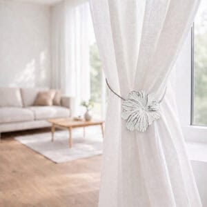 08 (living room lifestyle): Sheer white curtain held back by the flower tieback in a bright living room with a neutral sofa and wood coffee table softly blurred behind.
