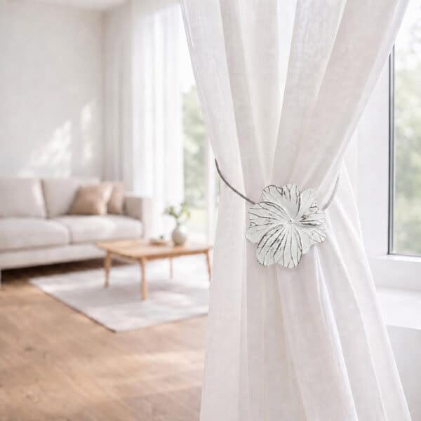 08 (living room lifestyle): Sheer white curtain held back by the flower tieback in a bright living room with a neutral sofa and wood coffee table softly blurred behind.