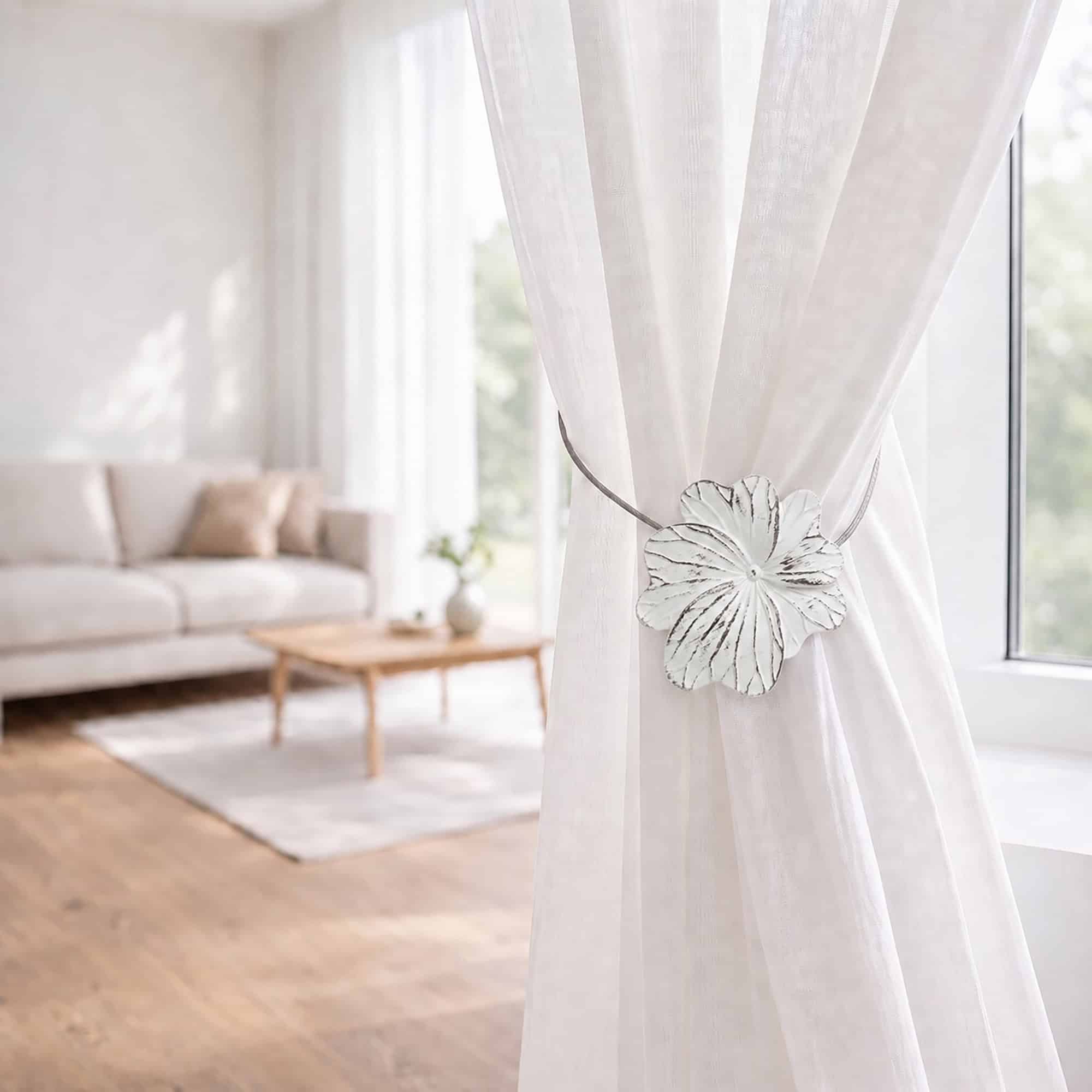 08 (living room lifestyle): Sheer white curtain held back by the flower tieback in a bright living room with a neutral sofa and wood coffee table softly blurred behind.