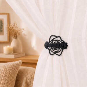 Close-up lifestyle view of a white sheer curtain gathered with a decorative black rose-shaped clip-on tieback in a cozy, warmly lit bedroom setting.