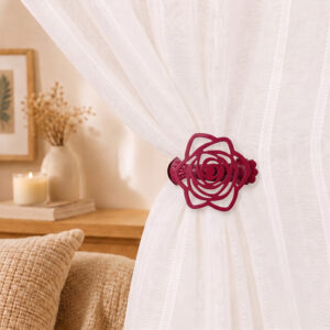 Close-up lifestyle view of a burgundy rose curtain holdback clip gathering a white sheer fabric panel in a cozy bedroom setting.