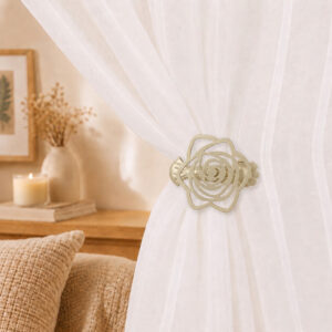 Close-up of a champagne gold rose-shaped clip holding together a white sheer curtain in a cozy, warm-toned living room setting.